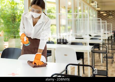 Portrait attraktive asiatische Kellnerin tragen Gesichtsmaske und Gesichtsschild Reinigungstisch mit Alkohol und nassen Tuch vor willkommenen Kunden. Neue normale Hygiene Stockfoto