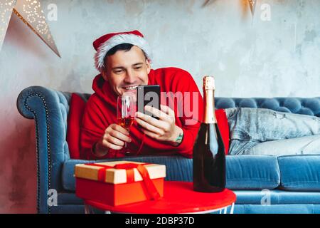 Junger schöner Mann in Weihnachtsmann Hut mit Glas Champagner Durchführung Live-Übertragung mit Freunden auf dem Smartphone. Fröhlicher Mann, der Weihnachten feiert Stockfoto
