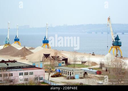 Dockkrane mit Industriegebäuden und großen Sandhaufen In der Nähe der Wolga in Russland Stockfoto