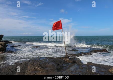 Rote Fahne auf einem hölzernen Fahnenmast fliegt an der Küste des Indischen Ozeans. Warnschild vor Gefahr durch große Leistungsschalter. Felsklippe am heiligen balinesischen Tempel Tanah Lot. Stockfoto