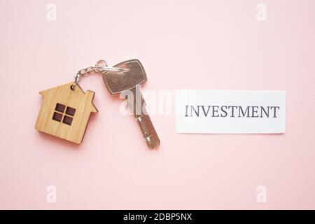 Holzhausspielzeug und silberner Schlüssel auf hellrosa Hintergrund mit Phrase Quote Investment. Hypothek, Hauskauf, Verkauf, Investition, Miete, Realtorkonzept Stockfoto