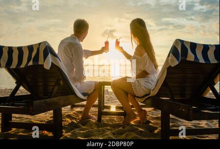 Junges Paar, das Cocktails am Strand während des Sonnenuntergangs auf Deck trinkt Stühle für Urlaub oder Flitterwochen in den Tropen Stockfoto