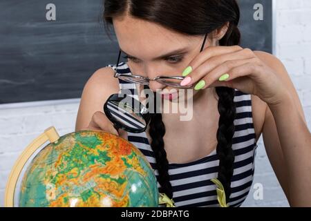 Trotz der dicken Gläser auf der Brille schaut ein Teenager durch eine große Lupe auf einen kleinen Globus. Interessante Geographie Lektion über unseren Planeten. Stockfoto