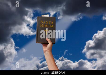 Arm in die Luft gehoben eine Hand nach oben und hält die Heilige Bibel in Spanisch Santa Biblia. Lichtstrahl, der durch dramatische, verschwommene Wolken kommt. S Stockfoto