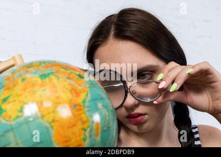 Trotz der dicken Gläser auf der Brille schaut ein Teenager durch eine große Lupe auf einen kleinen Globus. Interessante Geographie Lektion über unseren Planeten. Stockfoto