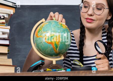 Trotz der dicken Gläser auf der Brille schaut ein Teenager durch eine große Lupe auf einen kleinen Globus. Interessante Geographie Lektion über unseren Planeten. Stockfoto