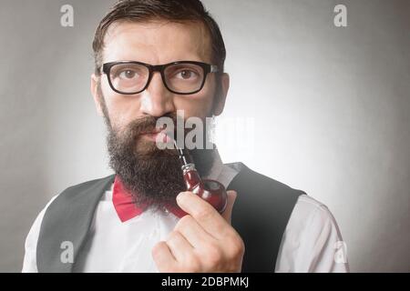 Vorderansicht Porträt von bärtigen eleganten Mann mittleren Alters mit Brillen, die Tabakpfeife auf grauem Hintergrund raucht. Stockfoto