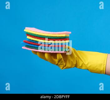 Hand in gelber Schutzlatex-Handschuh für die Reinigung eines Hauses hält einen Stapel Schwämme für die Reinigung, blauer Hintergrund Stockfoto