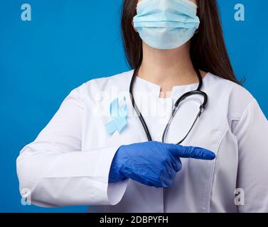 Arzt in einer sterilen Einwegmaske und einem weißen Mantel steht auf blauem Hintergrund und zeigt mit dem Zeigefinger zur Seite, ein blaues Seidenband in t Stockfoto