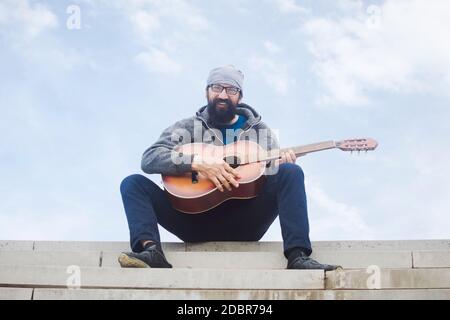 Der fröhlich lächelnde bärtige Mann spielt Gitarre und sitzt auf einer Treppe im Hintergrund des Himmels. Stockfoto