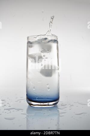 Ein Eiswürfel, der in ein Glas Wasser fällt und einen Spritzer erzeugt. Das Glas hat eine blaue Farbe an der Basis mit Spritzwasser rund um das Getränk. Stockfoto