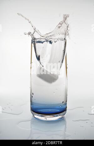 Ein Eiswürfel, der in ein Glas Wasser fällt und einen Spritzer erzeugt. Das blau getönte Glas spiegelt sich in den Wasserspritzern unter dem Getränk wider. Stockfoto