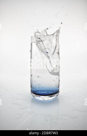 Ein Eiswürfel, der in ein Glas Wasser fällt und einen Spritzer erzeugt. Das Glas hat einen blauen Farbton am Boden und ist vor einem weißen Hintergrund. Stockfoto