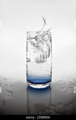 Ein Eiswürfel, der in ein Glas Wasser fällt und einen Spritzer erzeugt. Eine Spiegelung des Glases im Wasserbecken um das Getränk herum. Stockfoto