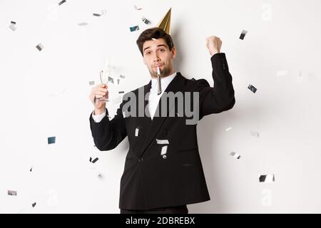 Schöner Party-Kerl im schwarzen Anzug Spaß haben, feiern neues Jahr, weht Pfeife und trinken Champagner, während Konfetti fallen, glücklich stehen Stockfoto