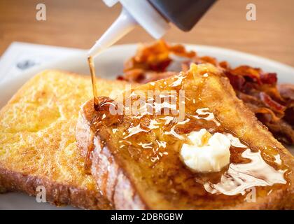 Zwei Stücke French Toast auf einem weißen Teller mit Speck verschwommen im Hintergrund. Daub Butter schmilzt auf, während Ahornsirup gegossen wird. Phot Stockfoto