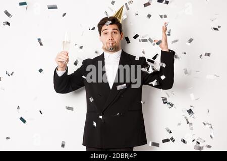 Schöner Party-Kerl im schwarzen Anzug Spaß haben, feiern neues Jahr, weht Pfeife und trinken Champagner, während Konfetti fallen, wünschen glücklich Stockfoto