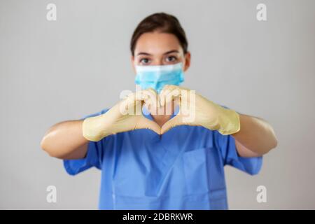 Junge Krankenschwester trägt eine Schutzmaske und Einweg-Latex-Handschuhe in einem Studio macht eine Herzform mit ihren Händen. Stockfoto