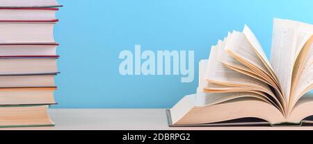 Ein großes, dickes Hardcover-Buch liegt auf einem hellen Holztisch. Das Buch ist offen, neben einem Stapel geschlossener Bücher, und auf der Rückseite ist ein blauer Hintergrund Stockfoto