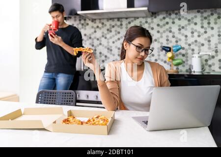 Asiatische Frau arbeiten von zu Hause in der Küche und nehmen Pizza, während asiatische Mann trinken Kaffee im Hintergrund, während Stadt Sperre von Coronavirus covid-19 Stockfoto