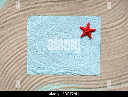 Leeres blaues Blatt Papier mit roten Seestern auf hellem Meersand. Textur aus leichtem Sand. Konzept für Strandurlaub. Sommerkonzept. Flach liegend, Draufsicht, Kopie Stockfoto