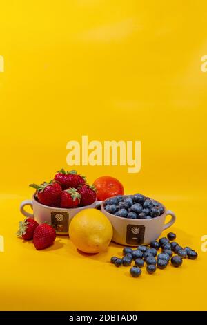 Erdbeeren und Heidelbeeren in Keramikbecher neben einer Zitrone und Mandarine vor gelbem Hintergrund Stockfoto