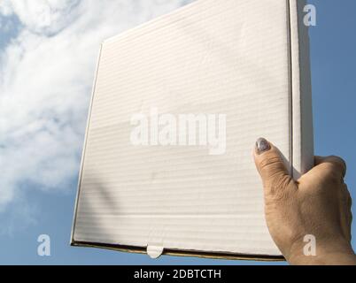Eine Frauenhand hält eine weiße Pappschachtel mit einer Kopie des Raumes und des Platzes für Text auf einem blauen Himmel Hintergrund, Schablone, mockup. Stockfoto