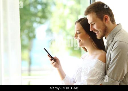 Seitenansicht eines Paares, das zu Hause neben dem Fenster auf dem Smartphone stand Stockfoto