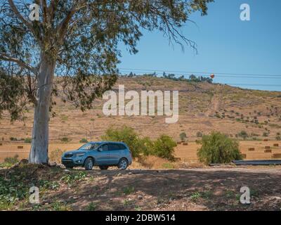Gray SUV off-road vehicle parked under a Eucalyptus tree shade on a dirt road. The landscape of mountains and wheat fields with hay bales in the count Stockfoto