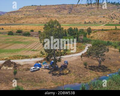 Landschaft des Landes Jordantals in Israel mit einer Gruppe von Menschen, die campen. Geländewagen und Zelte unter einem Baum in der Nähe eines Flusses. Berge und w Stockfoto