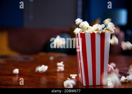 Popcorn fliegen aus Karton. Rot-weiß gestreifter Popcorn-Eimer mit fliegendem Popcorn im Wohnzimmer, Film- oder Kinokonzept Food oder Snac Stockfoto