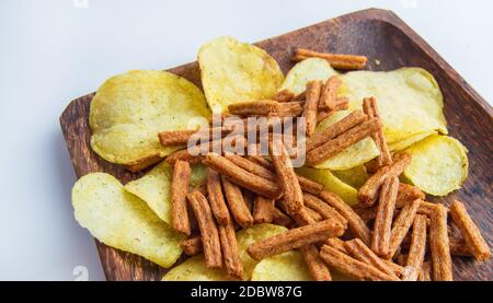 Leckere Kartoffelchips und gesalzene Roggenbrot-Cracker auf einem dunklen Holzteller, auf weißem Hintergrund. Kalorienreicher Snack. Stockfoto