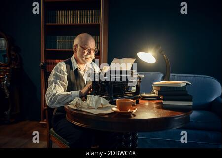 Älterer Schriftsteller arbeitet an Vintage-Schreibmaschine in seinem Heimbüro. Alter Mann in Gläsern schreibt Literaturroman im Raum mit Rauch, Inspiration Stockfoto