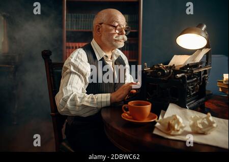 Älterer Schriftsteller arbeitet an Vintage-Schreibmaschine in seinem Heimbüro. Alter Mann in Gläsern schreibt Literaturroman im Raum mit Rauch, Inspiration Stockfoto