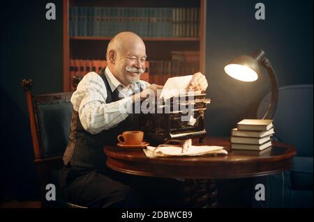 Lächelnder älterer Schriftsteller arbeitet an Retro-Schreibmaschine im Heimbüro. Alter Mann in Gläsern schreibt Literaturroman im Raum mit Rauch, Inspiration Stockfoto