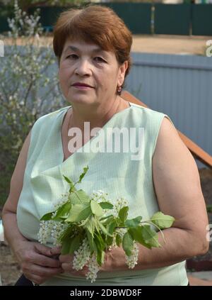 Portrait von 65 Jahre ältere Frau in ärmelloser Bluse mit neutralem Gesichtsausdruck sitzend und mit dem Frühlingsstrauß der blühenden Zweige haltend Stockfoto