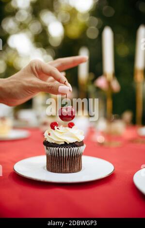 Tischdekoration, Tee-Party, Frau schmücken Kuchen mit frischer Kirsche. Luxus-Besteck auf roter Tischdecke, Geschirr im Freien. Hochzeitsfeier auf summ Stockfoto