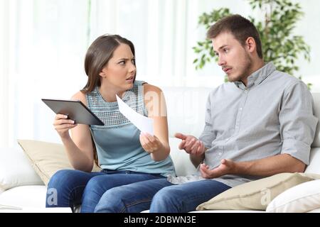 Verärgerte Frau mit Tablet, das dem Ehemann die Quittung zeigt, der zu Hause auf dem Sofa im Wohnzimmer sitzt Stockfoto