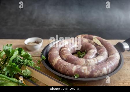 Hausgemachte rohe Schweinewurst wird auf einem Holztisch in einer gusseisernen Pfanne zum Braten zubereitet. Neben Gemüse und Gewürzen. Speicherplatz kopieren Stockfoto