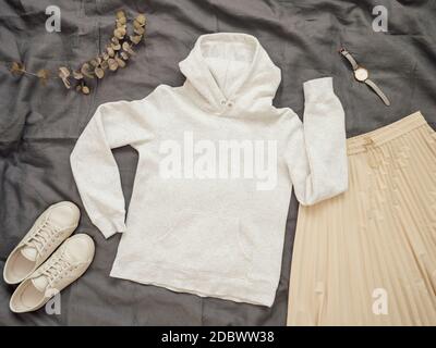 Modischer Damen-Look mit weißem leerem Hoodie, cremefarbenem Faltenrock und weißen Sneakers. Draufsicht auf weißen leeren Kapuzenpullover mit langen Ärmeln über grauem Bett Stockfoto