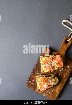 Gesunde offene Sanwiches mit dunklem Roggenbrot, Avocado, geräuchertem Lachs und Sprossen auf Holzschneidebrett auf grauem Hintergrund, Draufsicht, Kopierraum Stockfoto