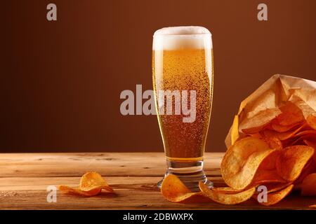 Schließen Sie ein Glas Lagerbier mit weißen Schaum und Blasen und Papier Tüte Kartoffelchips auf hölzernen Tisch über dunkelbraunen Hintergrund mit Kopie Raum Stockfoto