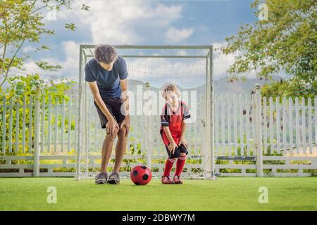 Kleiner niedlicher Junge in roter Fußballuniform und sein Trainer oder Vater spielt Fußball, Fußball auf dem Feld, im Freien. Aktives Kind macht Sport mit Stockfoto