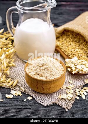 Mehl Hafer in der Schüssel, Milch in Krug, Haferflocken in Löffel auf Elop, Getreide in Beutel, Haferhalme gegen dunkle Holzbrett Stockfoto