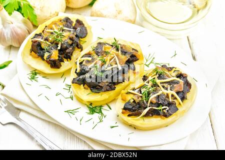 Kartoffeln gefüllt mit Pilzen, gebratenen Zwiebeln und Käse in einer Platte auf Serviette, Pflanzenöl im Dekanter, Petersilie, Knoblauch und einer Gabel auf dem Hintergrund Stockfoto