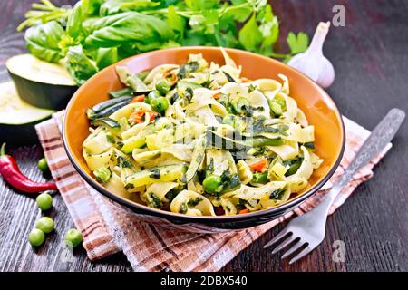 Tagliatelle Pasta mit Zucchini, grünen Erbsen, Spargelbohnen, Paprika und Spinat in einem Teller auf Handtuch, Knoblauch, Gabel und Basilikum auf dem Hintergrund Stockfoto