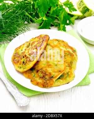 Fritten von Zucchini, Dill und Petersilie in einem Teller auf einem Handtuch, saure Sahne in Untertasse auf dem Hintergrund von hellen Holzbrett Stockfoto