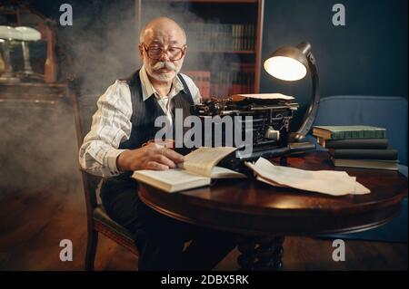 Älterer Schriftsteller arbeitet an Vintage-Schreibmaschine in seinem Heimbüro. Alter Mann in der Brille schreibt Literaturroman Stockfoto