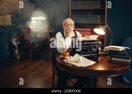 Älterer Schriftsteller arbeitet an Vintage-Schreibmaschine in seinem Heimbüro. Alter Mann in Gläsern schreibt Literaturroman im Raum mit Rauch Stockfoto