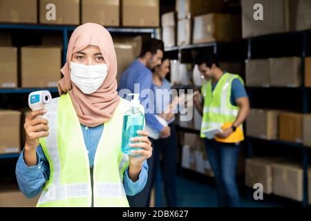 Asiatische Islam muslimische weibliche Lagerarbeiter mit Gesichtsmaske halten Hand Desinfektionsmittel und Infrarot-Thermometer. Für neue normale Hygiene Leben im Geschäft wareho Stockfoto
